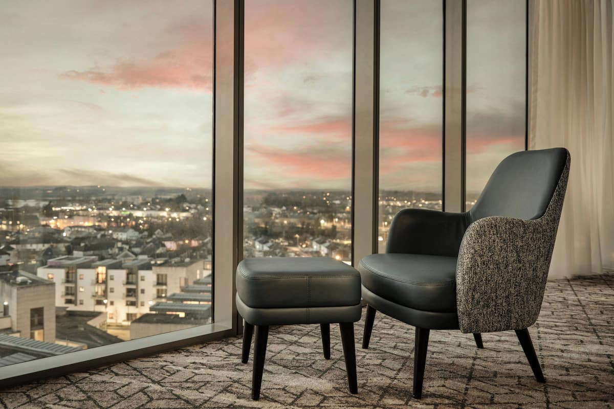 A room with a view of a cityscape through large windows, a black leather chair, a grey woven ottoman, and a patterned carpet.