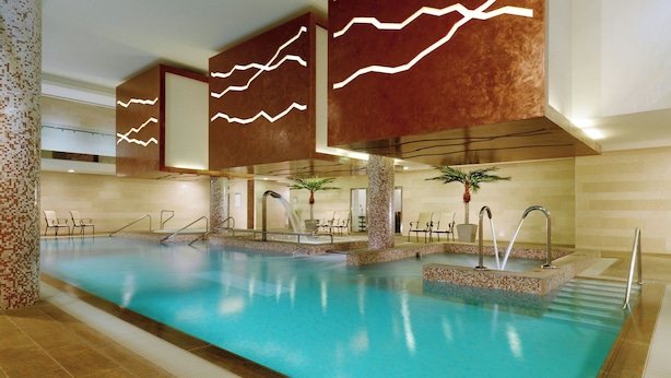 Una amplia piscina interior con agua cristalina, rodeada de sillones de descanso y palmeras, y diseños modernos en las paredes.