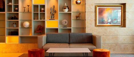 Un salon moderne avec un canapé gris, une table basse blanche et deux poufs rouges. On y trouve des étagères en verre où sont exposés divers objets décoratifs, ainsi qu’un tableau encadré accroché au mur.