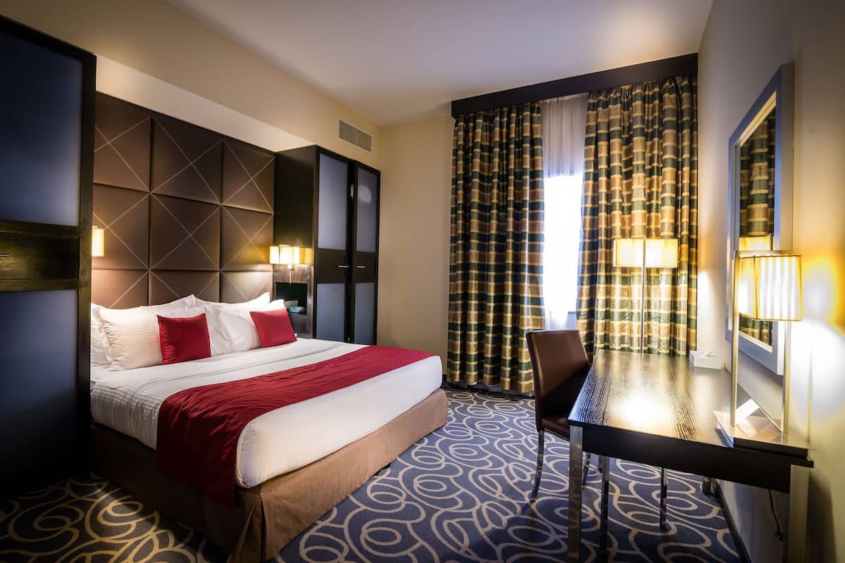 Ein Hotelzimmer mit einem großen Bett, einem Schreibtisch und einem Stuhl. Ein Fenster mit Vorhängen und eine Lampe auf dem Schreibtisch.