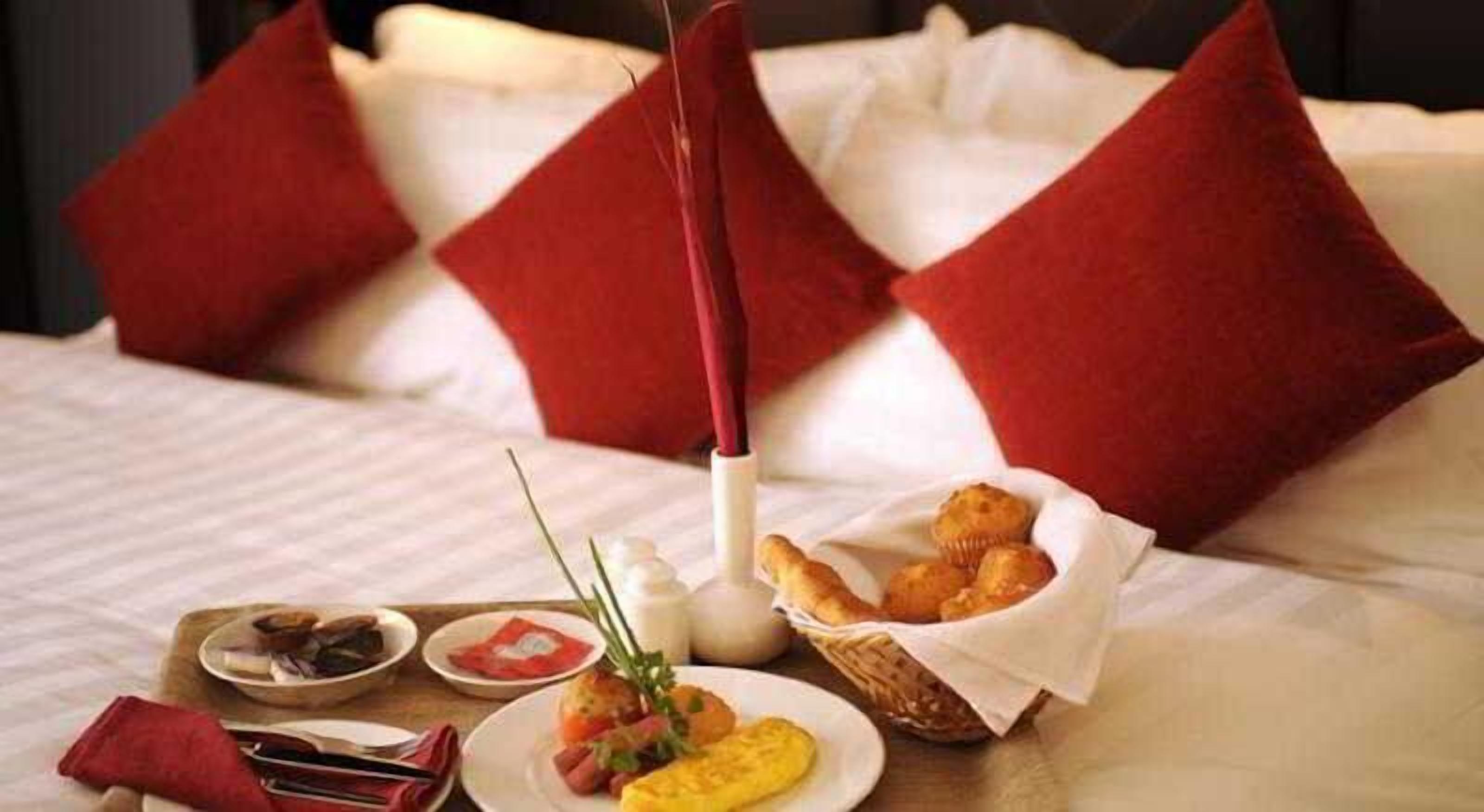 A neatly arranged breakfast table with a variety of food items, including eggs, pastries, and a bottle of milk, set on a bed with red pillows.