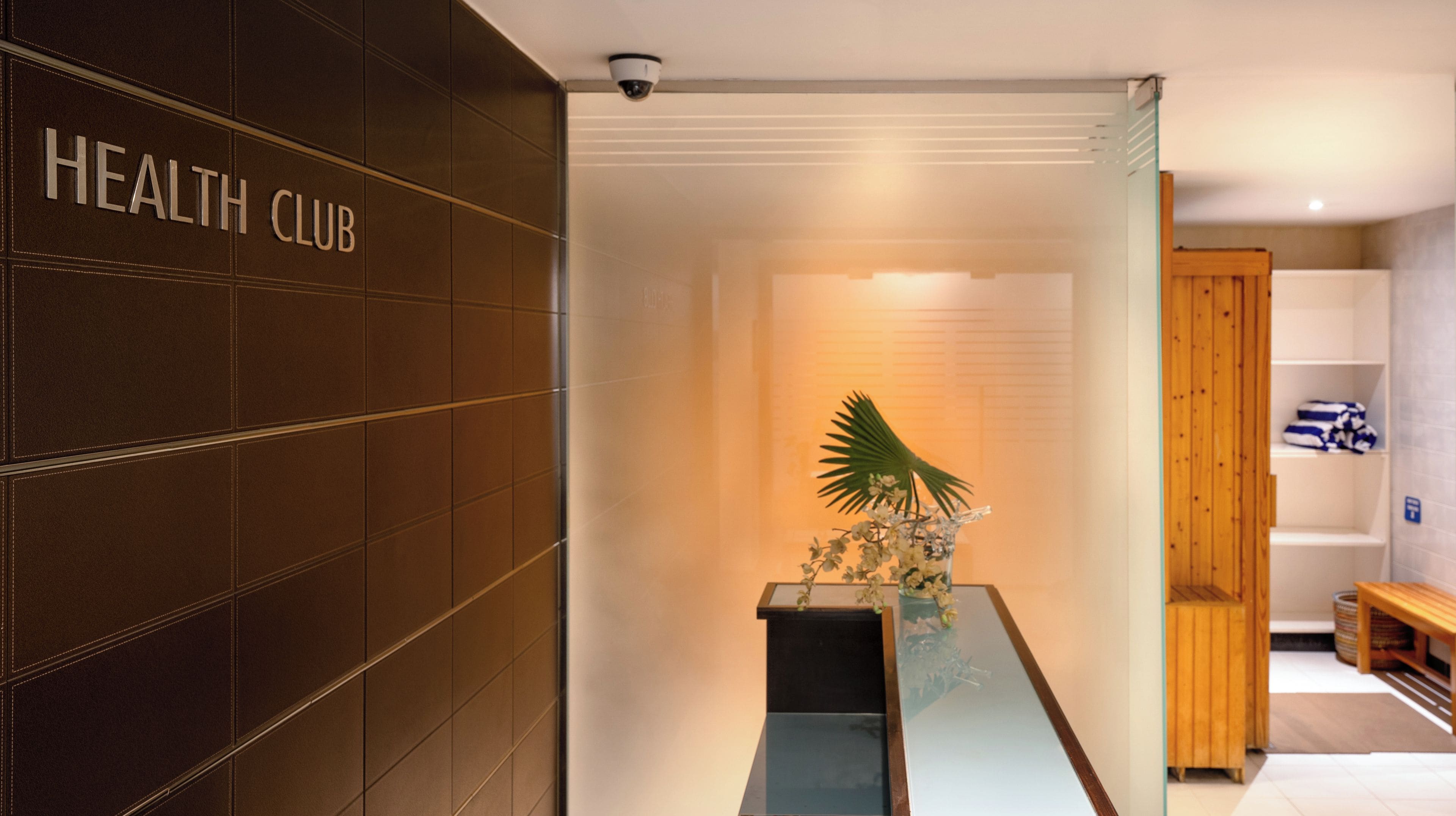 A reception area with a 'HEALTH CLUB' sign, a reception desk, a potted plant, and a wooden bench.