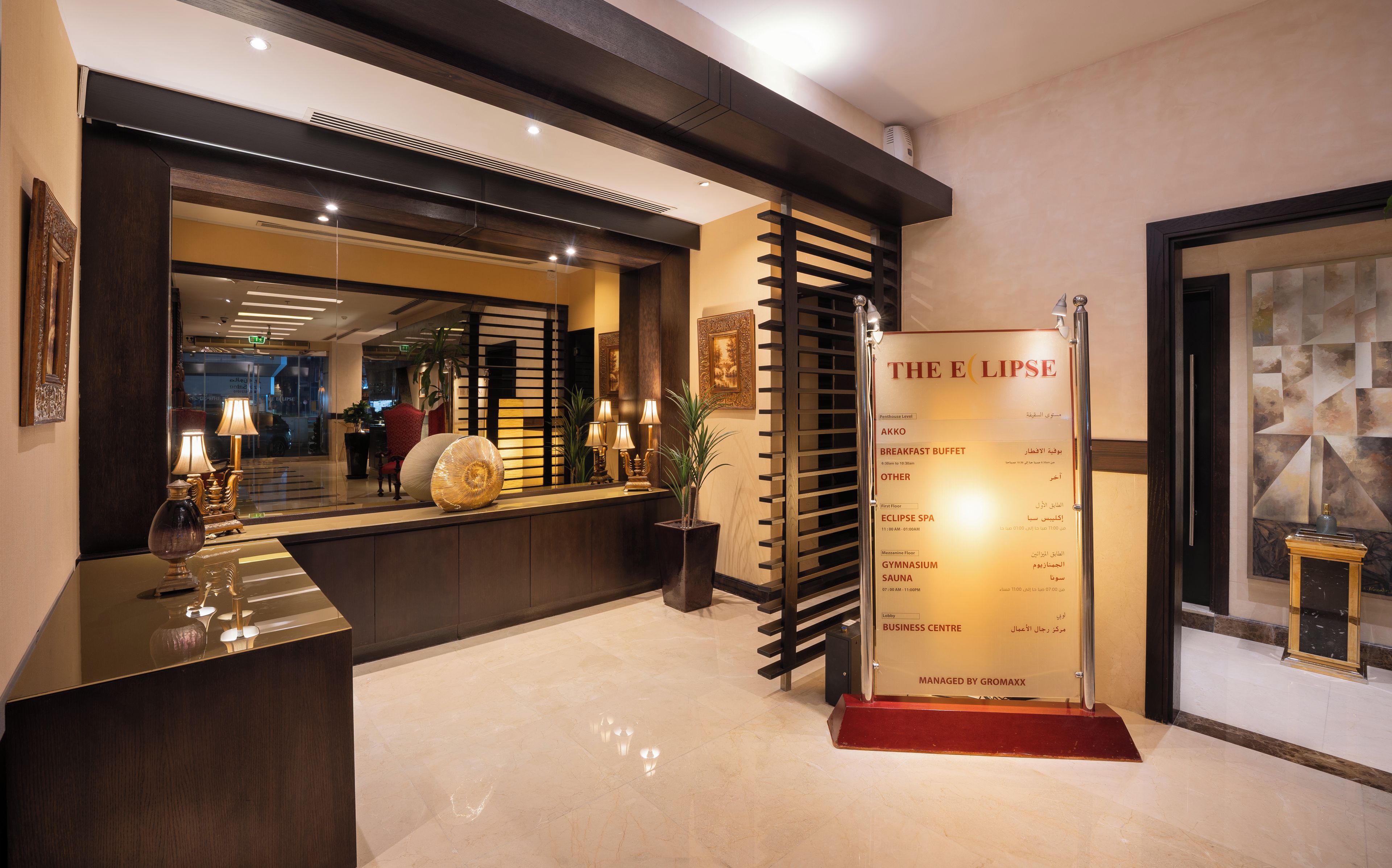 A reception area with a polished reception desk, a mirror, and a wall-mounted information board listing amenities such as a breakfast buffet and spa.