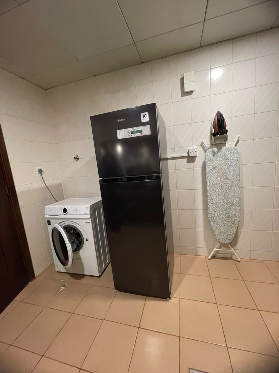 A compact kitchen with a black refrigerator, a washing machine, and a patterned ironing board.