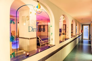 A hotel corridor with arched doorways, a reception desk, and a visible room with a bed and a nightstand.