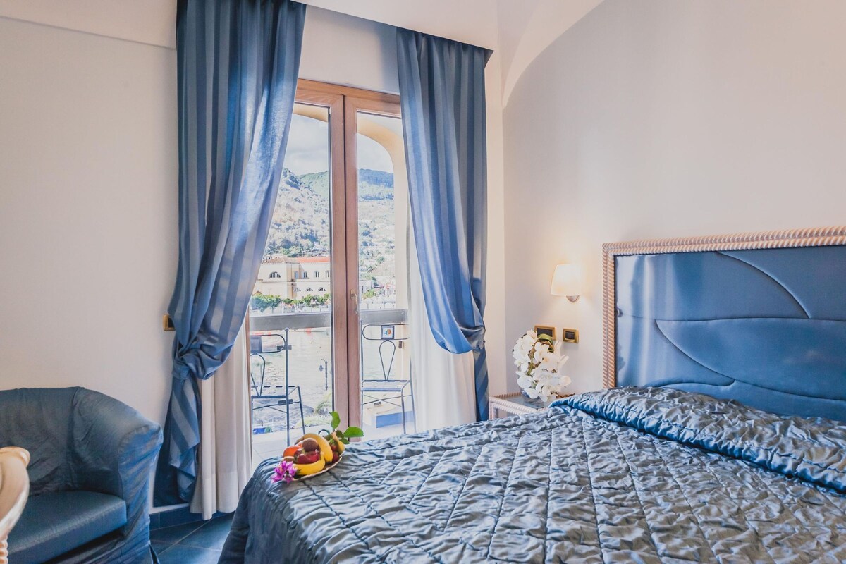 A hotel room with a bed, a chair, a balcony with curtains, and a view of buildings.