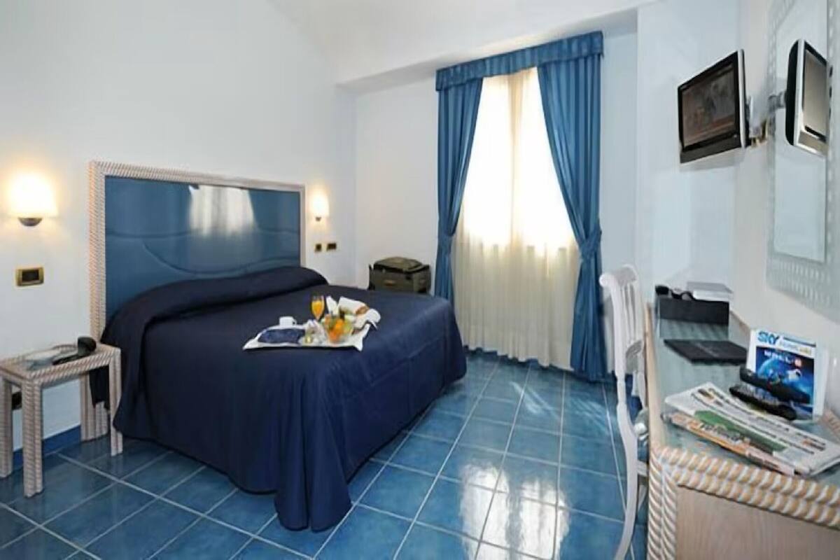 Ein Hotelzimmer mit einem Bett, einem kleinen Tisch mit einem Frühstücksbrettchen, einem Sessel, einem Schreibtisch mit einem Computer und einem Fenster mit Vorhängen.
