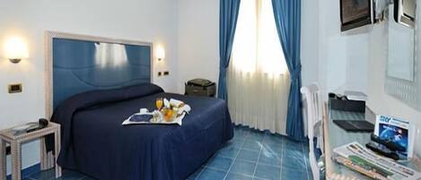 A hotel room with a bed, a small table with a tray of breakfast, a chair, a desk with a computer, and a window with curtains.