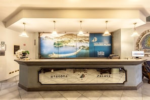 A hotel reception desk with a large window displaying a coastal view, a sign reading 'Hotel Aragona Palace,' and a reception area with a counter and overhead lighting.