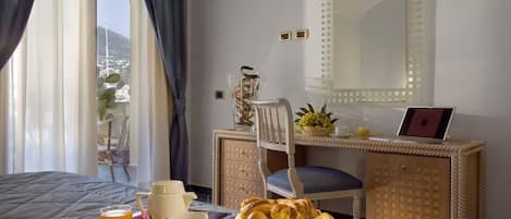 A hotel room with a bed, a desk with a laptop, a chair, a dining table with breakfast items, and a window with curtains.