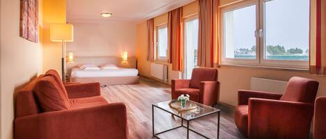 A hotel room with a bed, two red armchairs, a glass coffee table, and large windows with curtains.