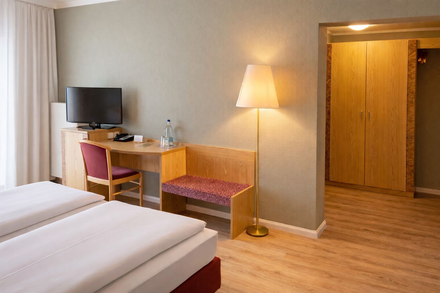 Ein Hotelzimmer mit zwei Betten, einem Schreibtisch mit Stuhl, einem Fernseher, einer Lampe und einem Kleiderschrank.