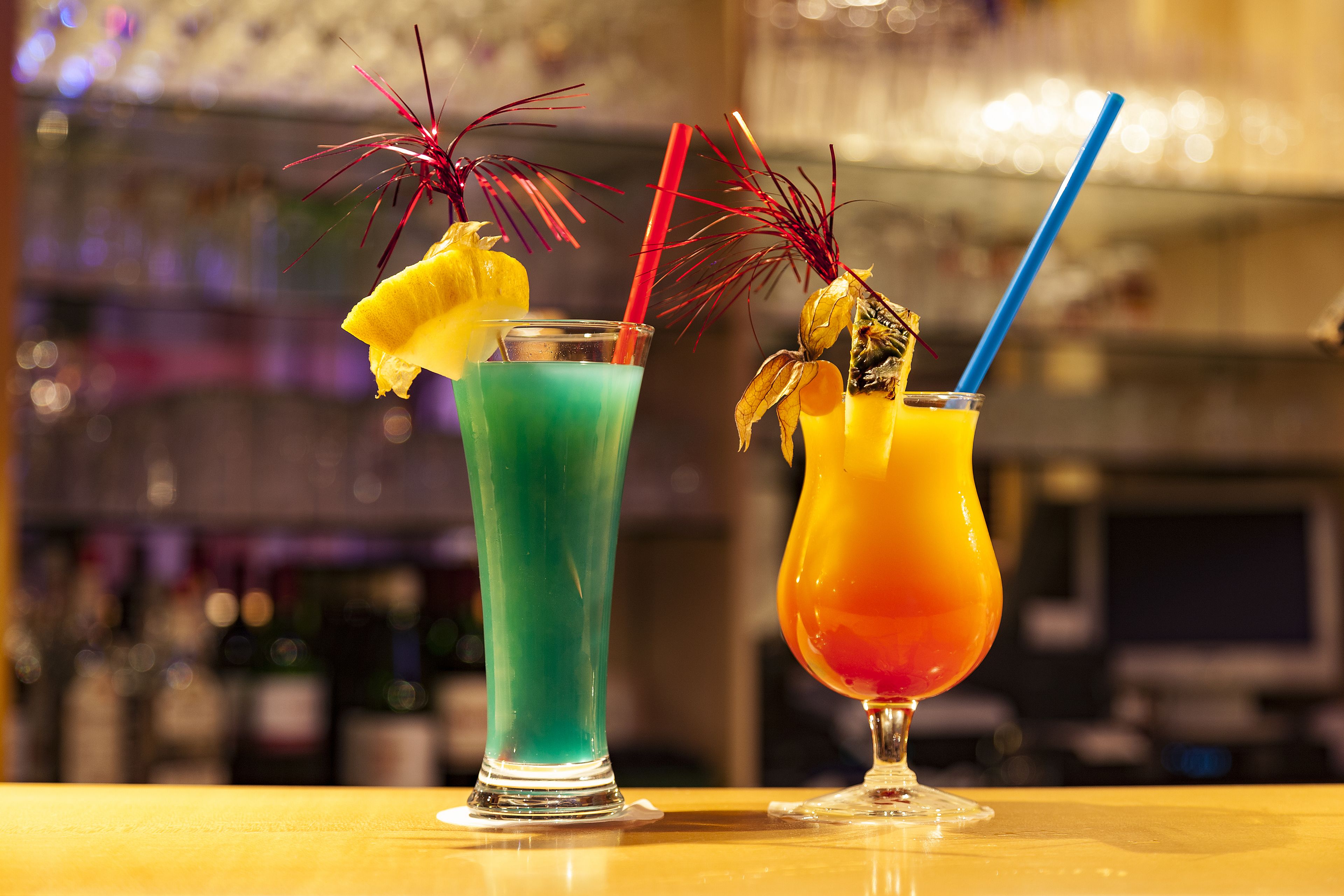 Two cocktails with straws and garnishes on a bar counter.