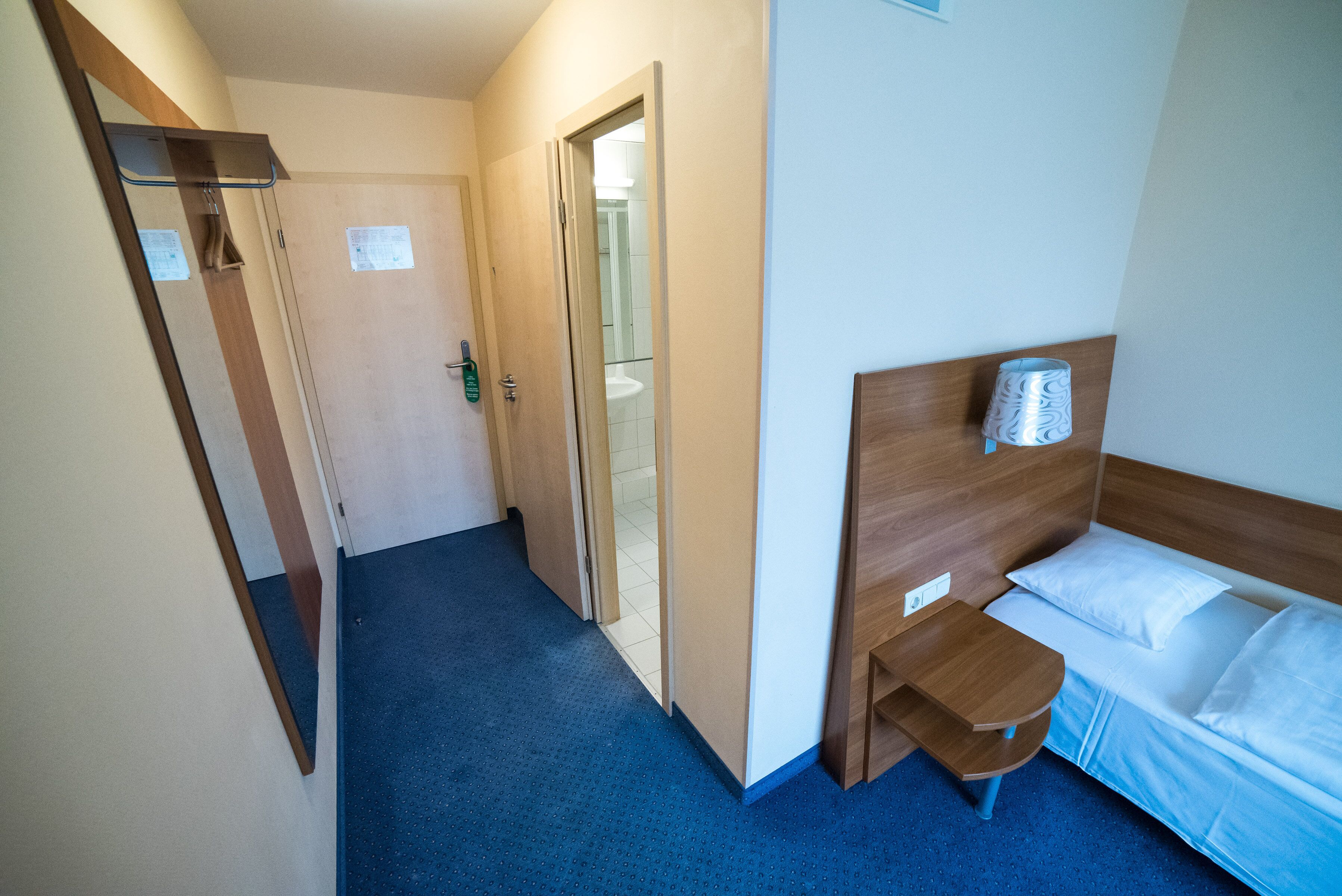 Una habitación de hotel con una cama individual, cabecera de madera, una mesita de noche pequeña y un espejo.