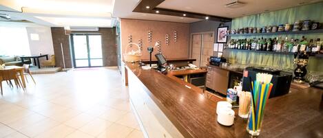 A hotel lobby with a reception desk, a bar area with shelves of bottles, and a seating area with chairs and a table.