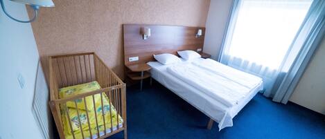 A hotel room with a wooden crib, a bed with white linens, a nightstand, and a lamp.