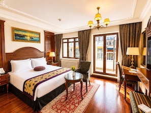 A hotel room with a large bed, a sitting area with a table and chairs, a view of the city through the window, and a chandelier. - GLK PREMIER Regency Suites & Spa - Special Class (Istanbul)