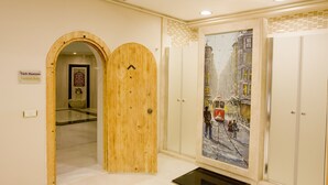 Un couloir avec une porte en bois portant l’inscription « bain turc », un mur recouvert de carreaux et un tableau représentant une rue enneigée.