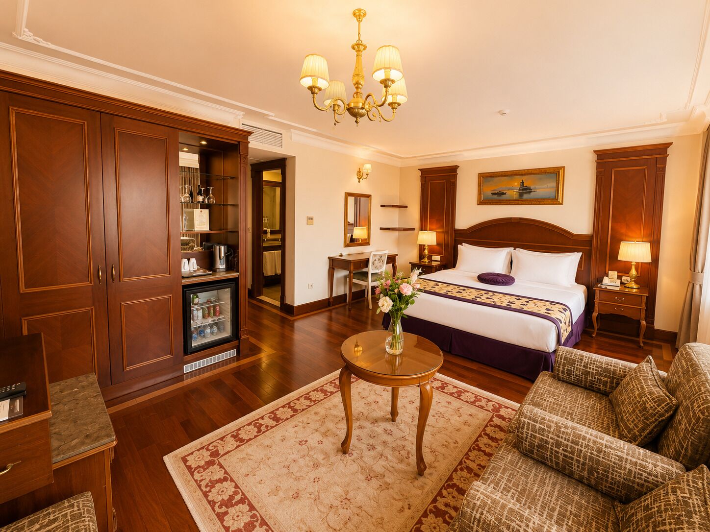 A hotel room with a bed, a desk, a refrigerator, a chandelier, and a view of the sea.