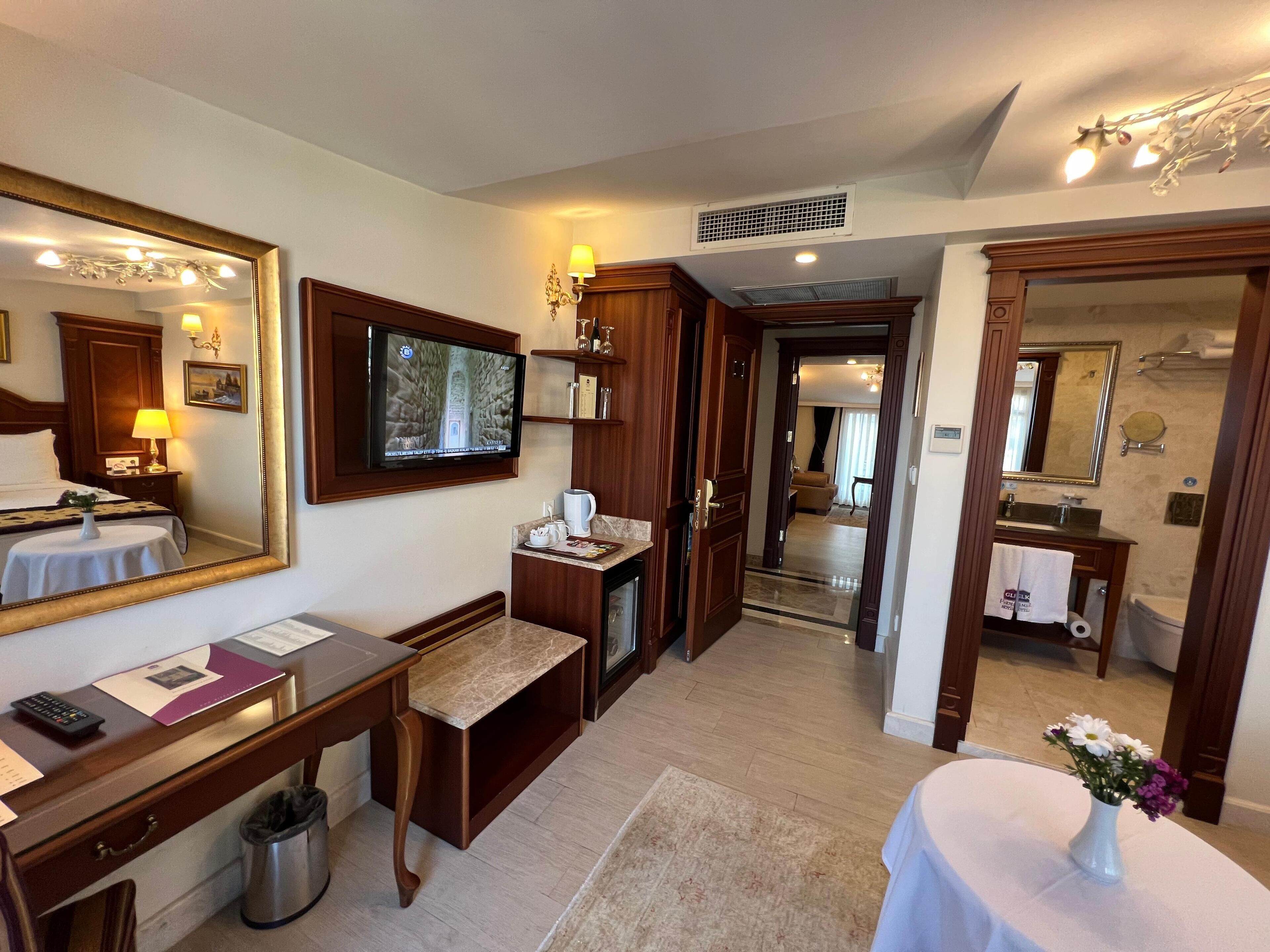 A hotel room with a large mirror, a television, a desk with a computer, a small table with a vase of flowers, and a view into the living area.
