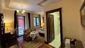 Une chambre d’hôtel comprenant un coin repas, un canapé, une petite table avec un vase, et une salle de bain visible à travers une porte ouverte.