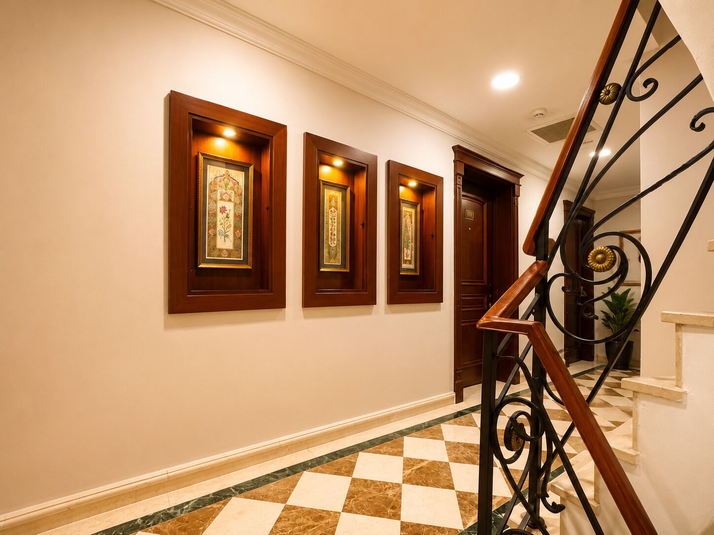 A hallway with a checkered floor, a wooden staircase with a metal railing, and three framed pictures on the wall.