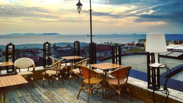 Une terrasse dotée de tables en bois et de chaises en osier, offrant une vue sur le paysage urbain de la côte, avec la mer et des îles lointaines en toile de fond.