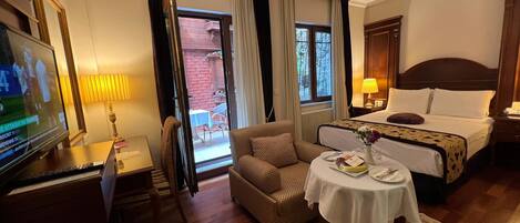 Quarto de hotel com cama, escrivaninha com televisão, cadeira, mesa com um vaso e vista para o exterior.