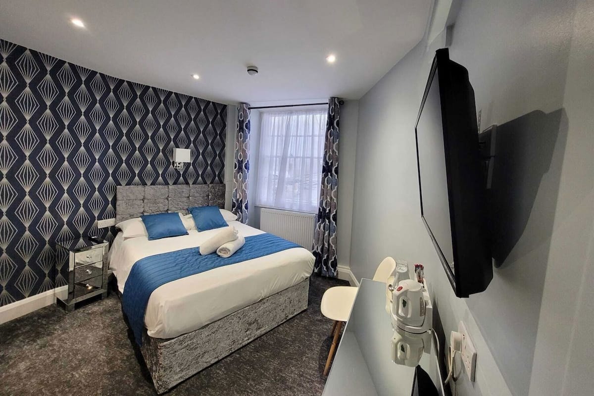 Una habitació d'hotel amb un llit, una taula de nit, una cadira, un televisor i una finestra amb cortines.