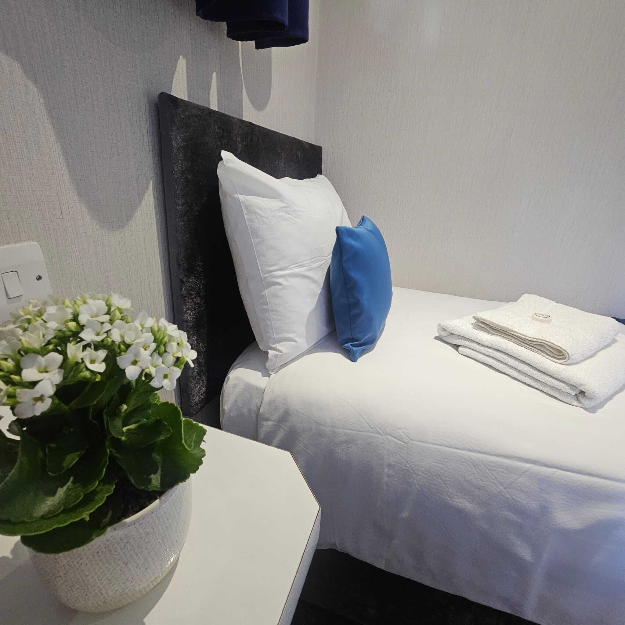 A neatly made bed with white bedding and a folded towel on the side. A blue pillow is placed on the bed, and a white pillow is on the headboard. There is a white flower pot with green leaves on a white table beside the bed.