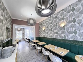 Un interno di ristorante dal design moderno, con lampade a sospensione a righe, carta da parati decorata e una varietà di posti a sedere, tra cui tavoli e banconi.