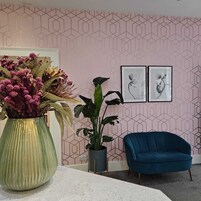 Una stanza con una parete a motivi geometrici, un vaso verde con fiori, una sedia blu e due quadri incorniciati.