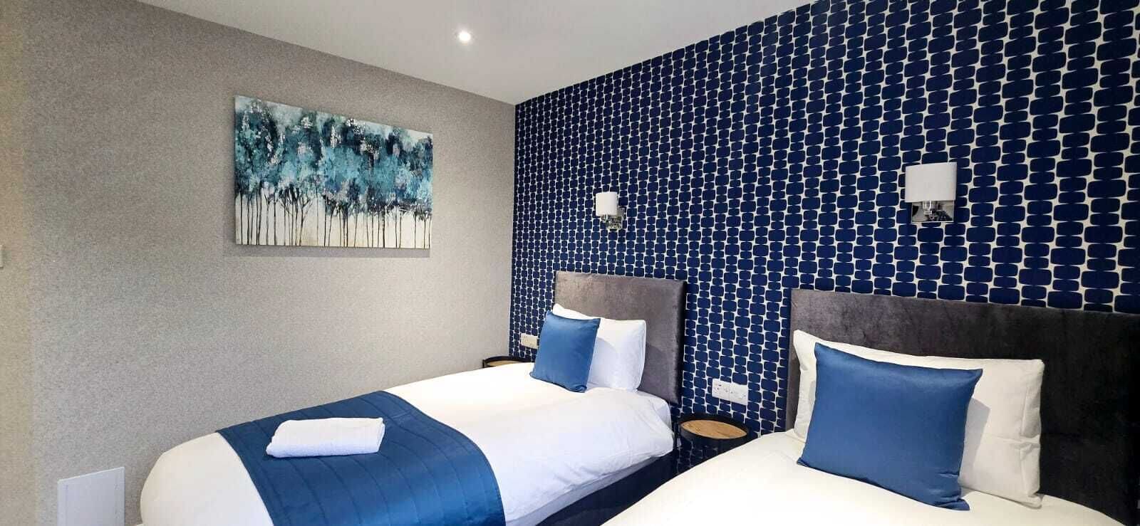 A hotel room with two beds, a blue and white color scheme, a large abstract painting on the left wall, and a patterned blue wall on the right.