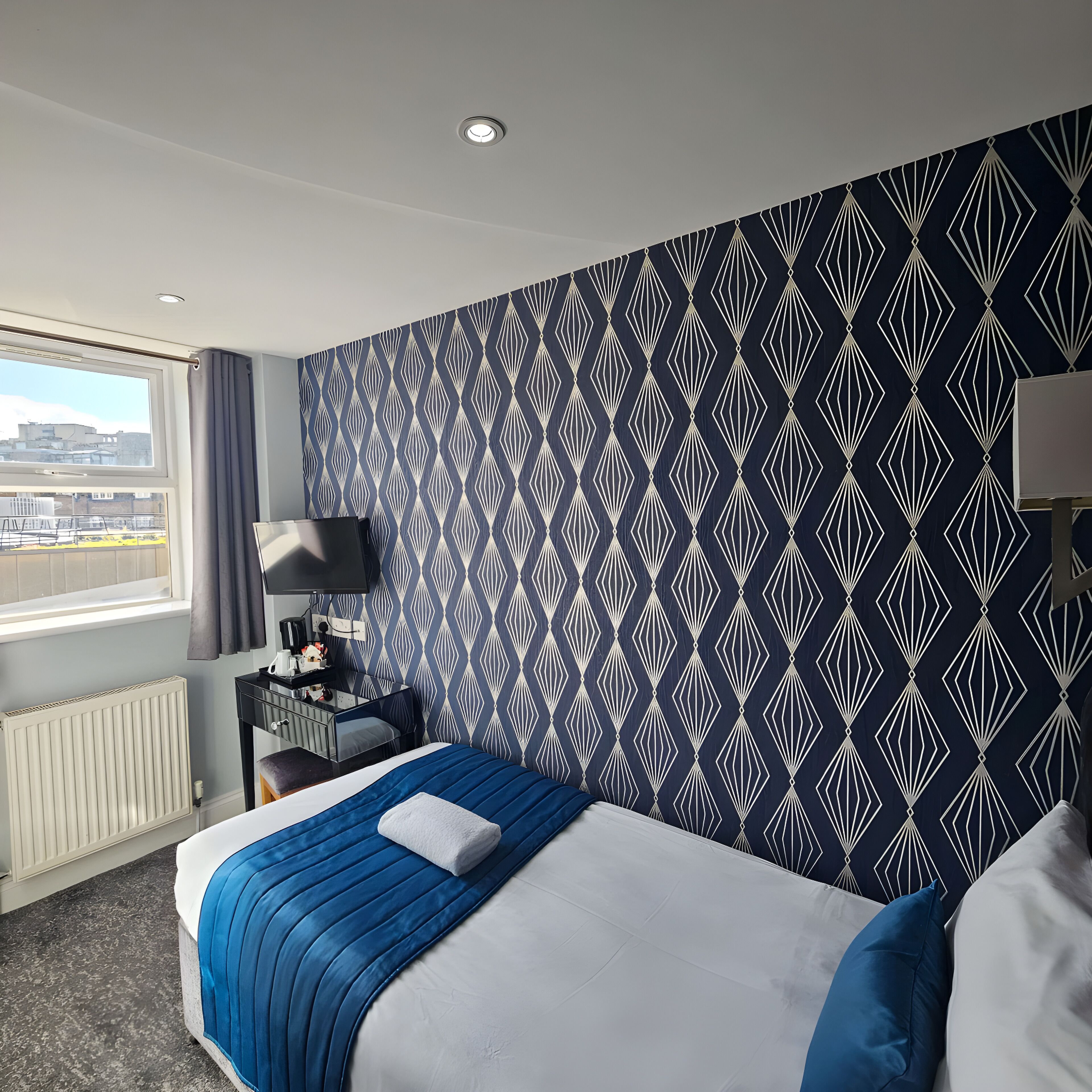 Habitación de hotel con una cama grande, un escritorio con lámpara, una ventana con vistas a edificios y una pared con diseño.