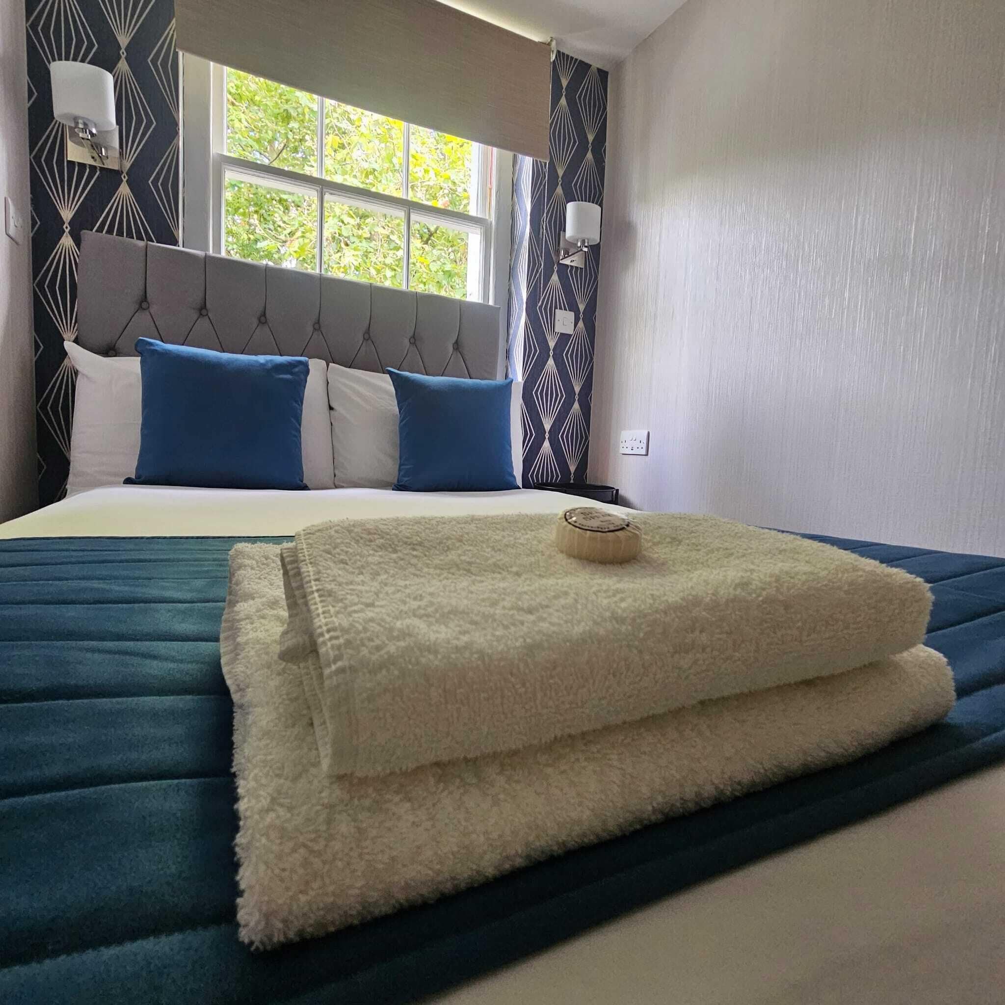 Una cama bien hecha con edredón azul y blanco, una manta blanca esponjosa y una toalla enrollada encima.