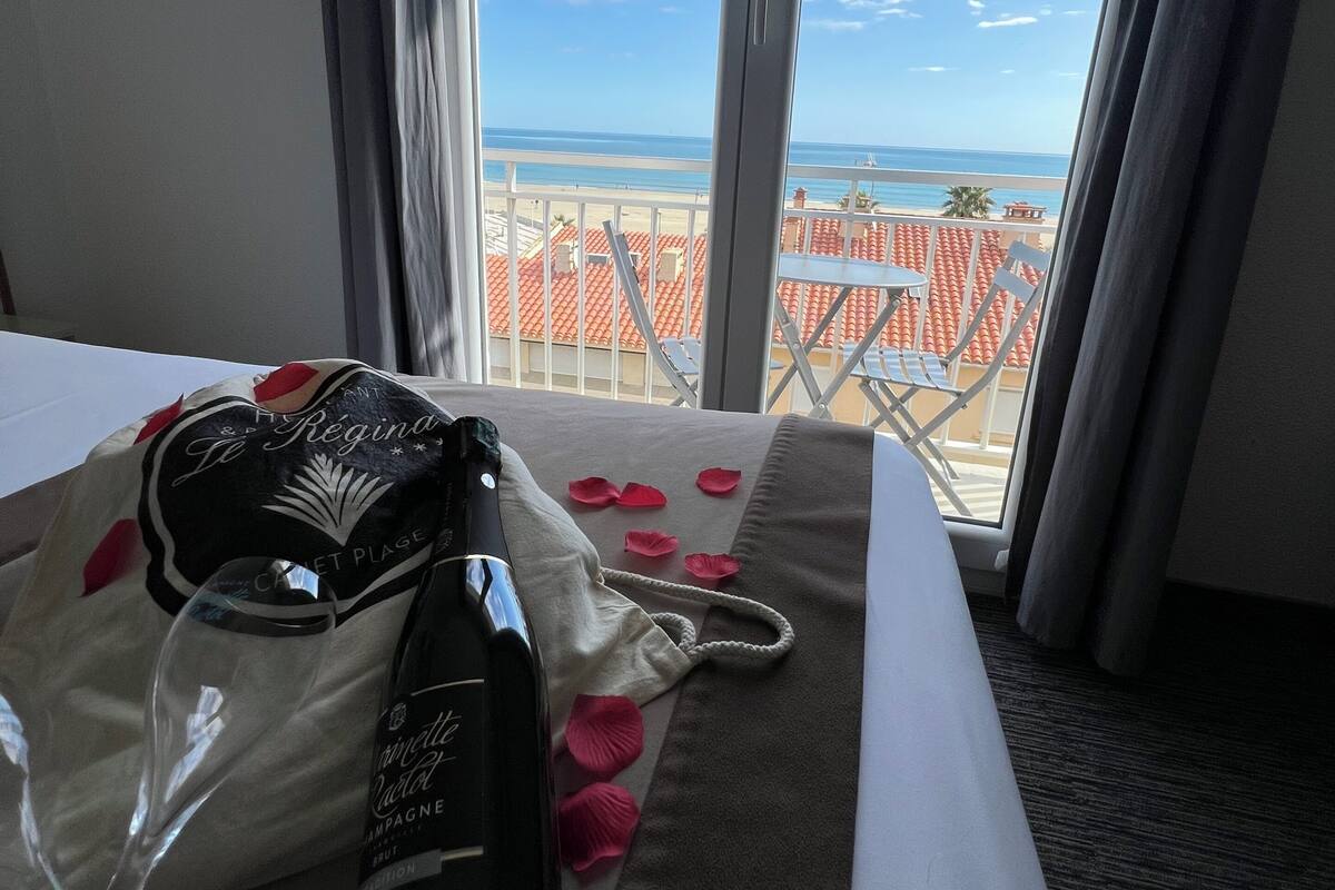 Habitación de hotel con cama, ventana con vista a la playa y al océano, y un mini bar con una botella y un vaso.