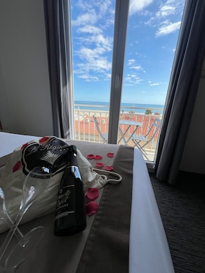 A hotel room with a bed, a window with a view of the beach and ocean, and a minibar with a bottle and a glass.