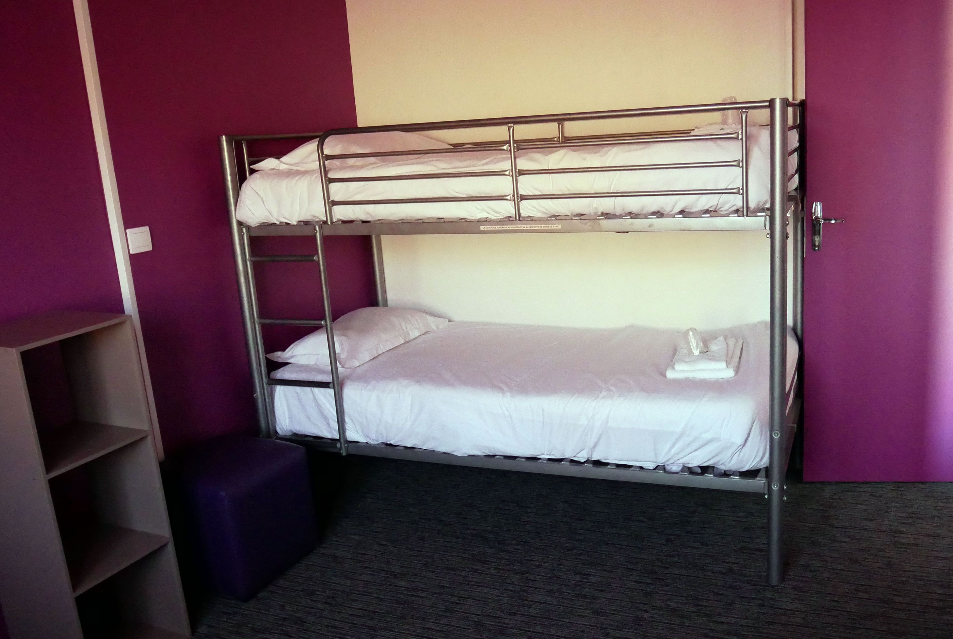 A bunk bed with a ladder, a purple ottoman, and a shelf unit.