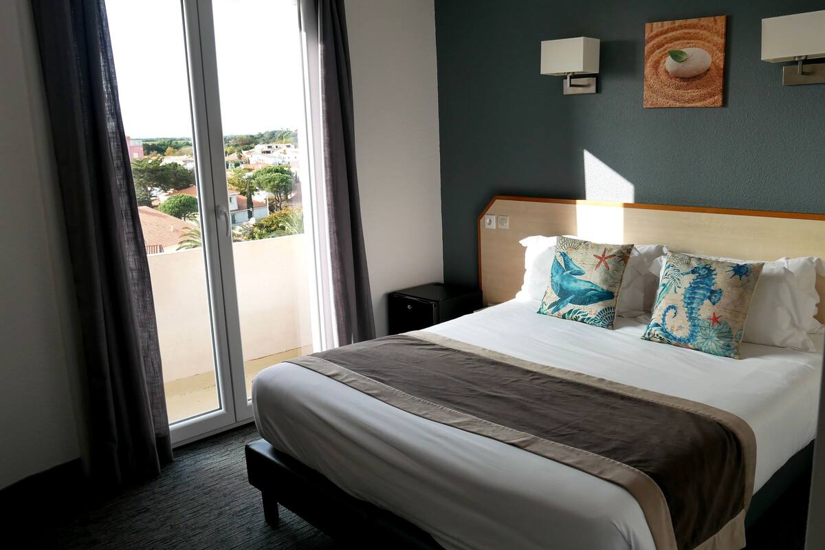 Una habitación de hotel con cama, mesita de noche, ventana con cortinas y vistas al exterior.