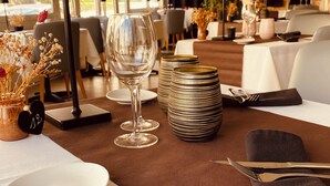A dining table set with plates, glasses, cutlery, and stacked plates in a well-lit room with large windows. - Le Regina (Canet-en-Roussillon)