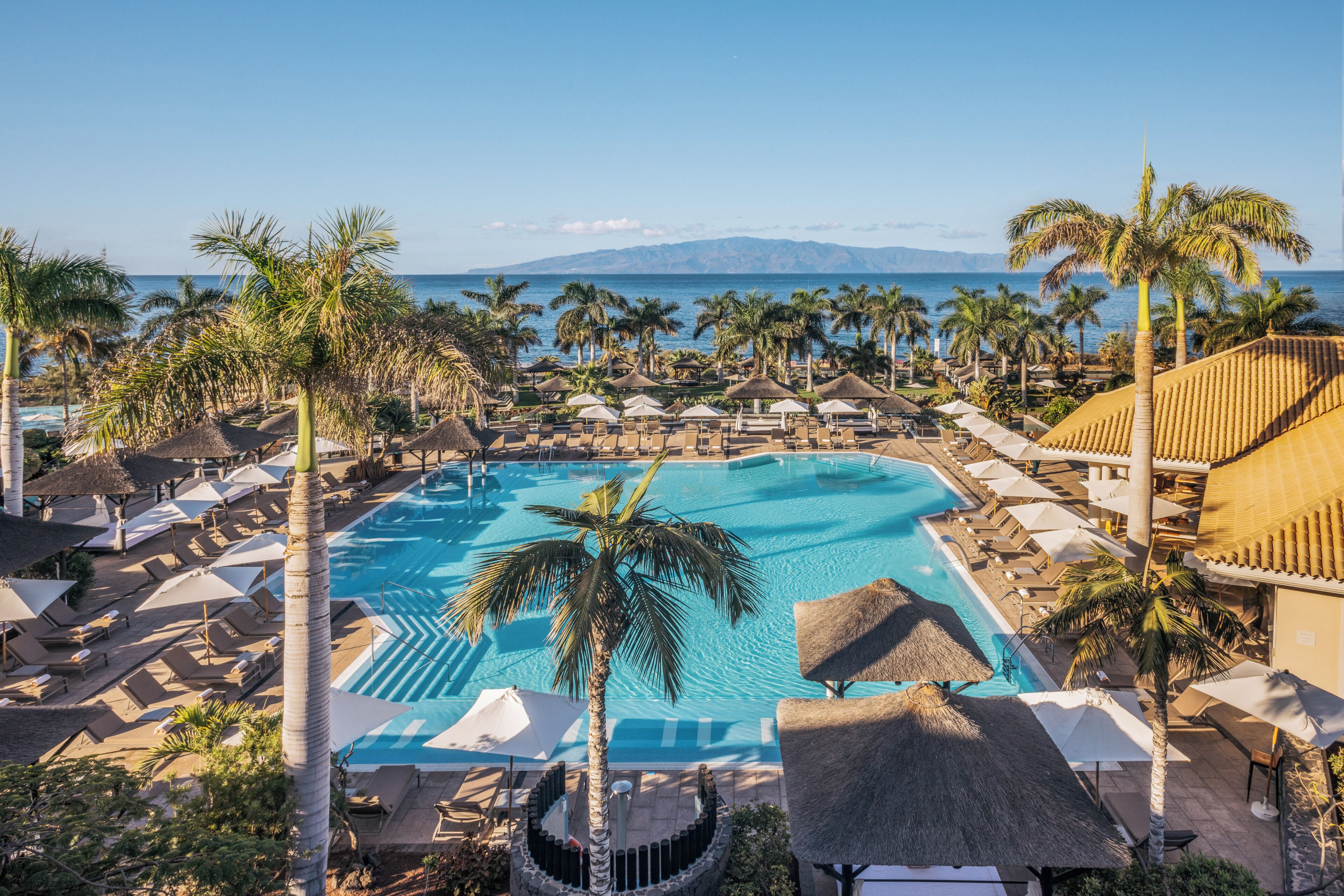 Una piscina de resort con palmeras, sillas de descanso y sombrillas.