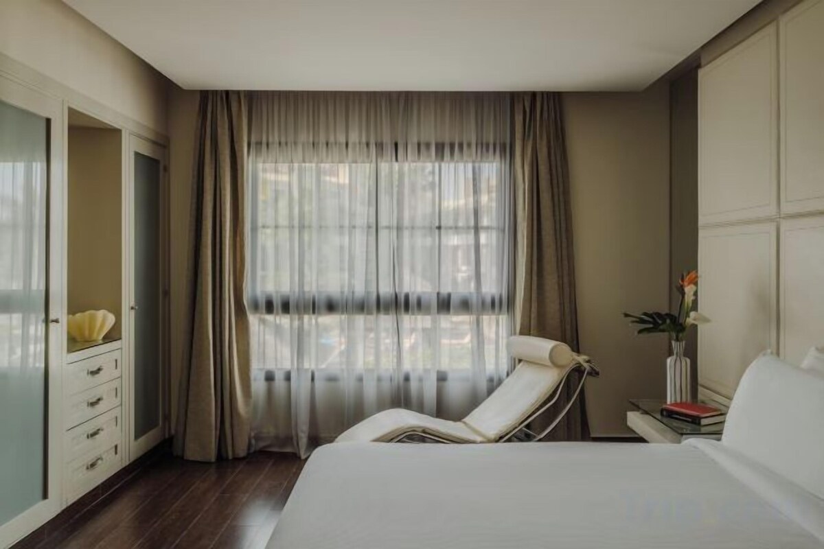 Une chambre d’hôtel avec un lit, une table de chevet ornée d’un vase de fleurs, une chaise, une armoire et une grande fenêtre avec des rideaux.