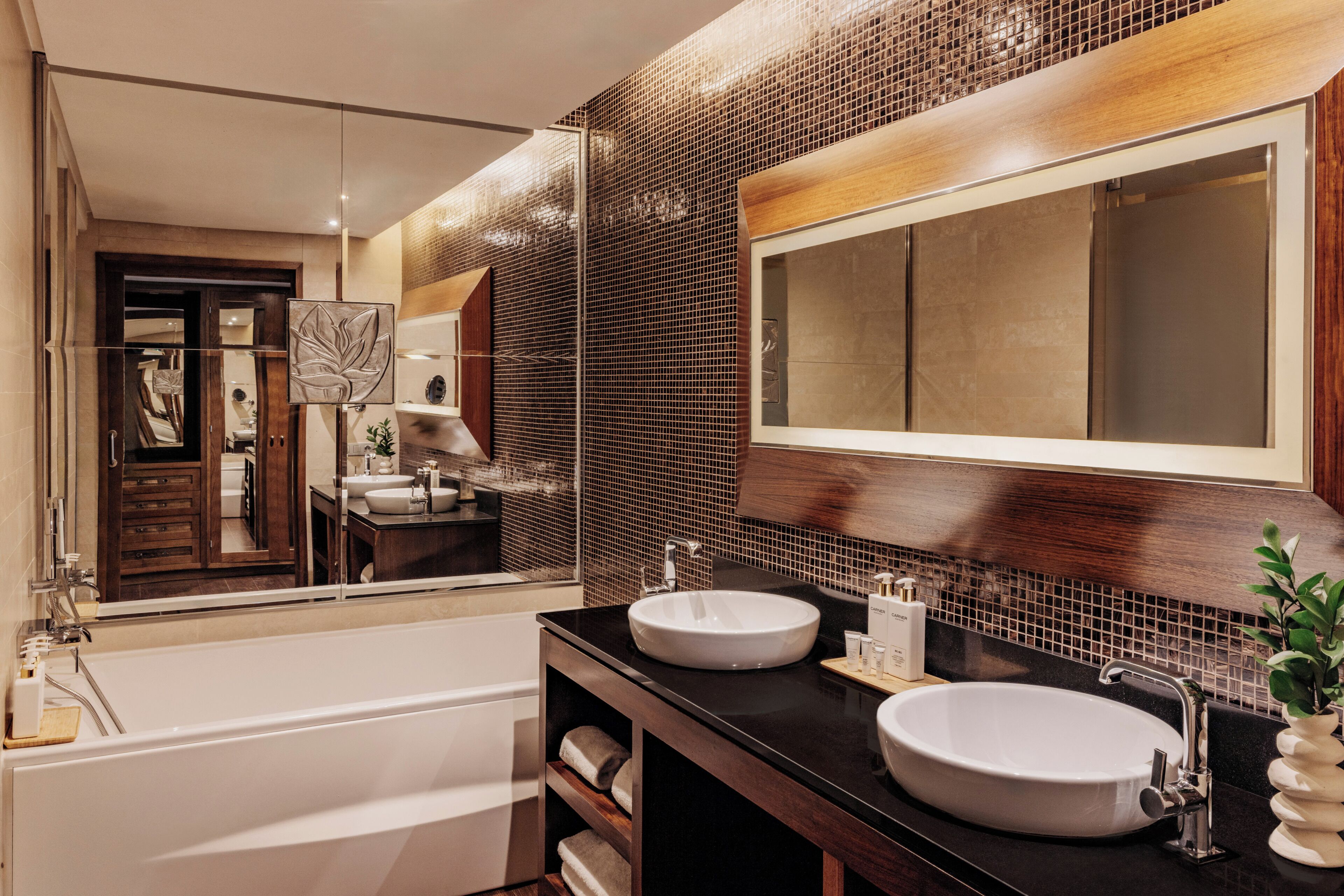 A modern bathroom with a bathtub, two sinks, a large mirror, and a decorative wall panel.