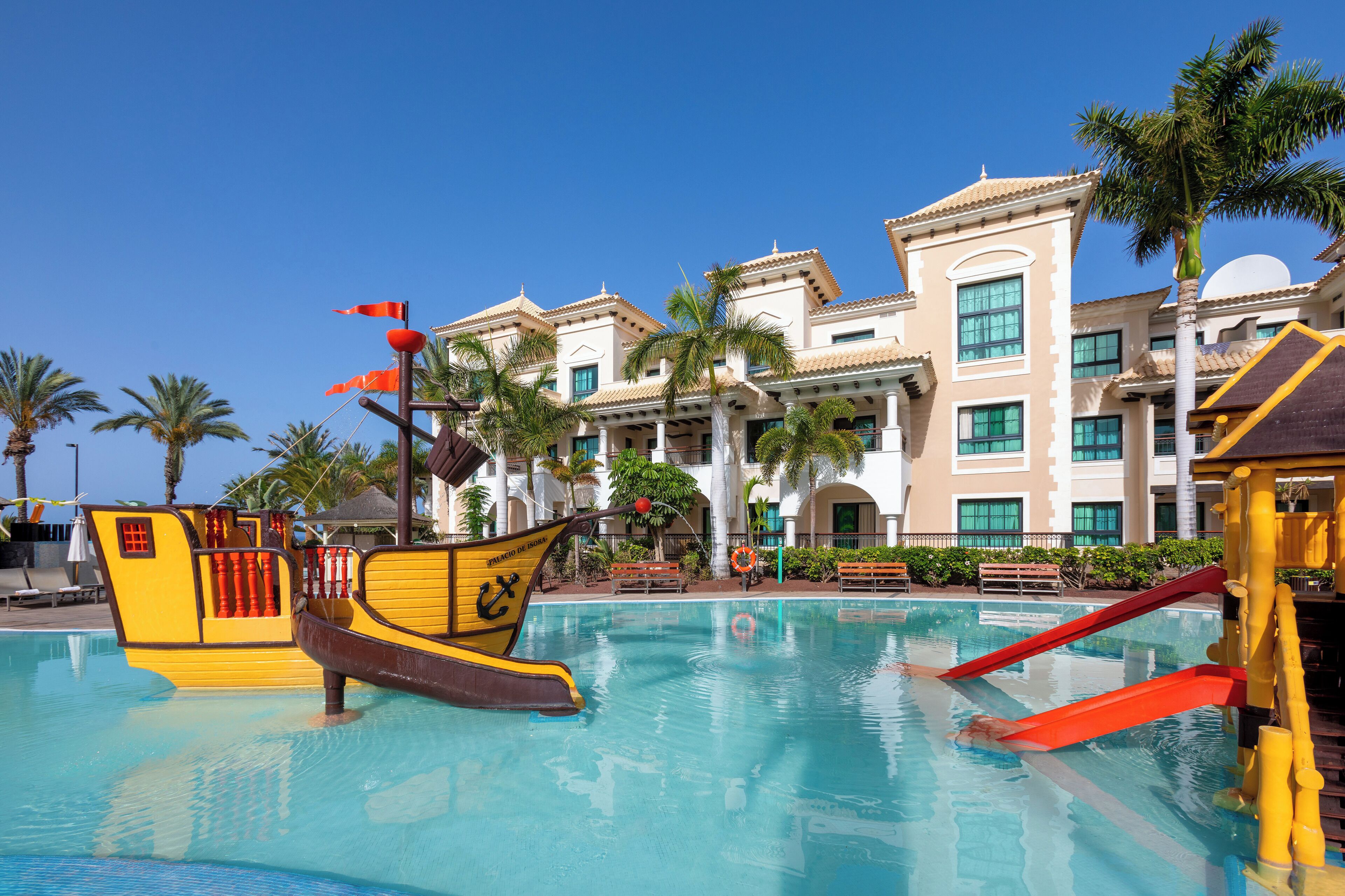 A swimming pool with a pirate ship decoration, palm trees, and a building with multiple windows and balconies.