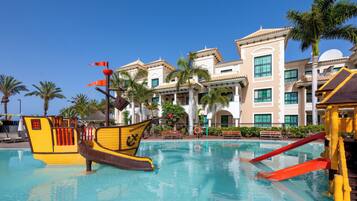 Una piscina con decoración de barco pirata, palmeras y un edificio con múltiples ventanas y balcones.