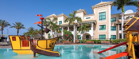 A swimming pool with a pirate ship decoration, palm trees, and a building with multiple windows and balconies.