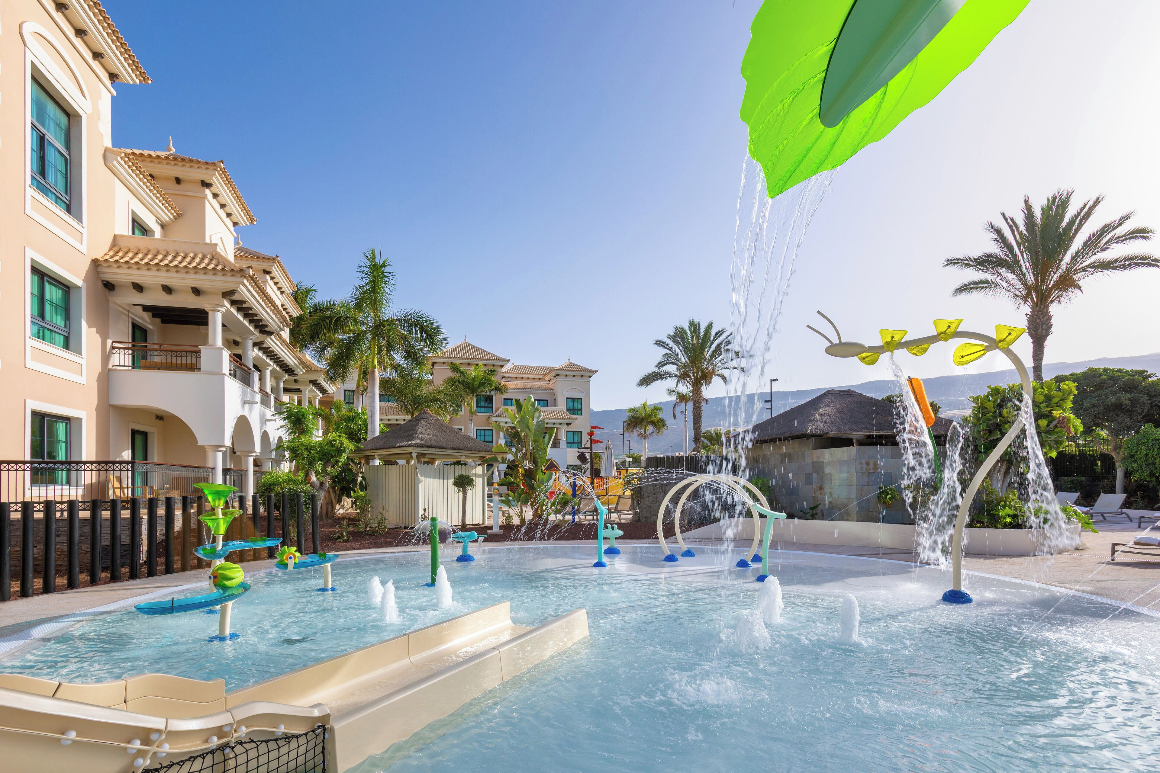 A pool area with multiple water features, including fountains and slides, surrounded by palm trees and buildings with balconies.