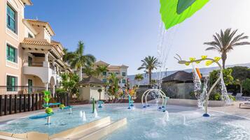 A pool area with multiple water features, including fountains and slides, surrounded by palm trees and buildings with balconies.
