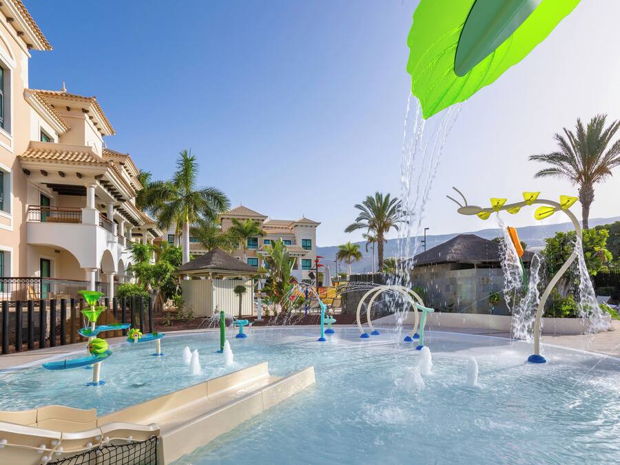Un espace piscine doté de plusieurs éléments aquatiques, notamment des fontaines et des toboggans, entouré de palmiers et de bâtiments avec des balcons.