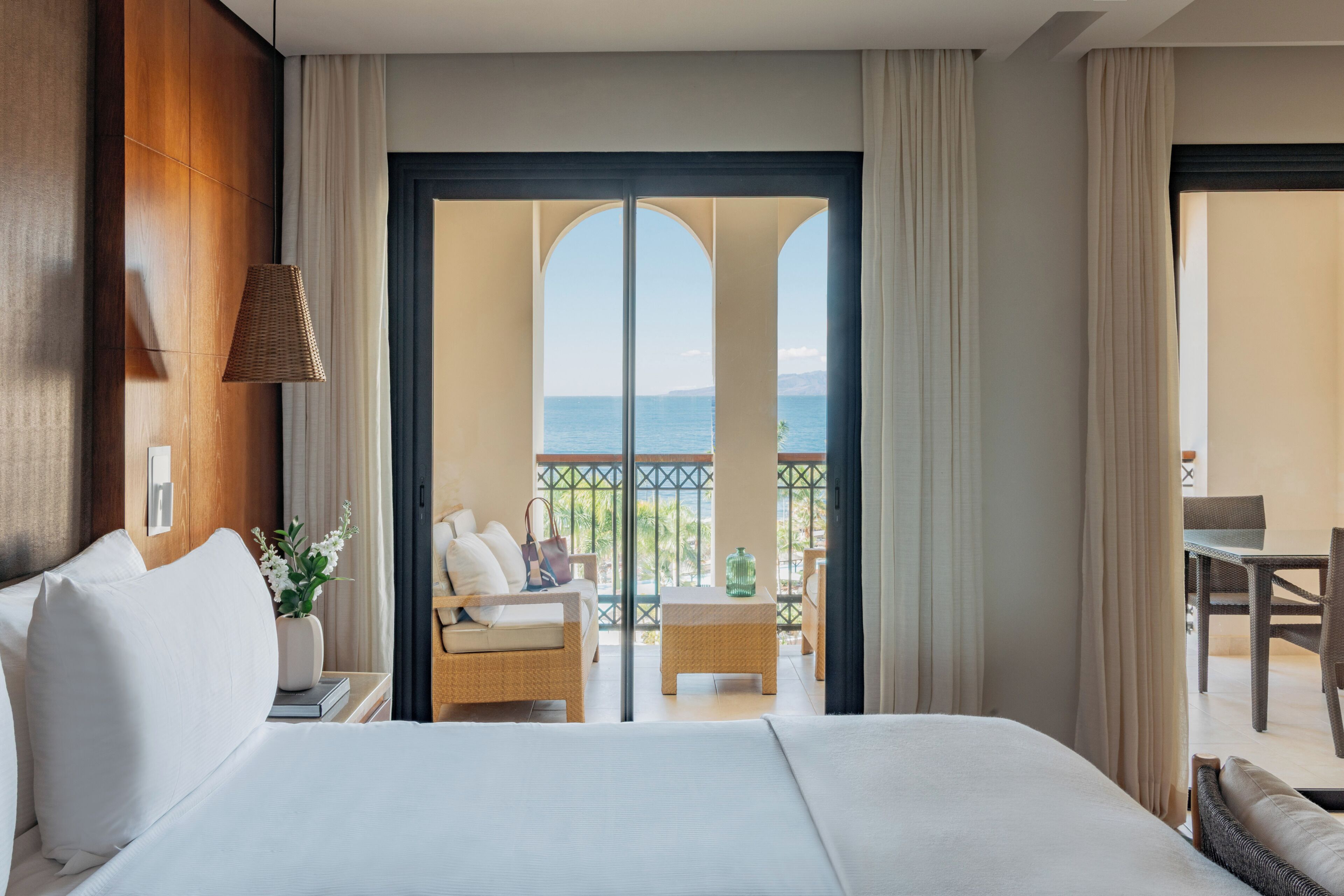 A hotel room with a bed, a small table with a vase, a chair, and a view of the sea through a glass door.